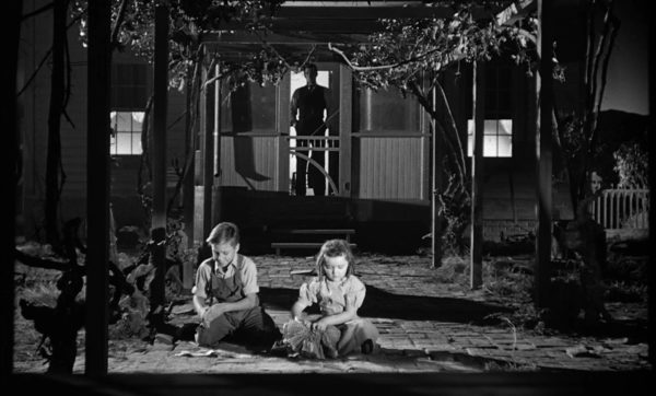 Robert Mitchum as Harry Powell, standing on the porch, looming ominously in the background as two children play in the foreground