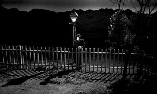 Robert Mitchum as Harry Powell leaning on a fence under a streetlamp