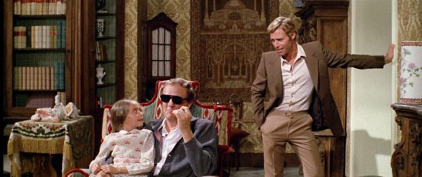 A blind man sits with a young girl in his lap while a blonde man stands nearby