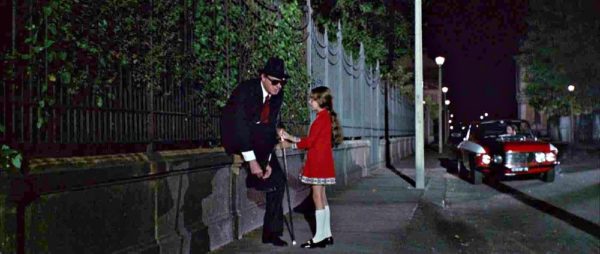 A blind man ties his shoe while a young girl in red dress holds his hand