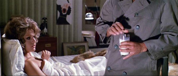 A woman sits upright in bed as the upper body of a man wearing a trench coat holds a glass of milk