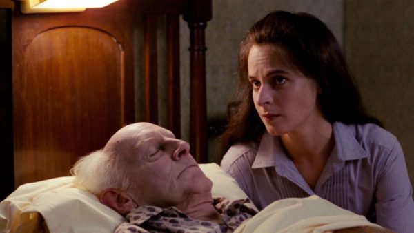 A young woman with dark brown hair sits on the left side of an old man lying in bed with his eyes closed