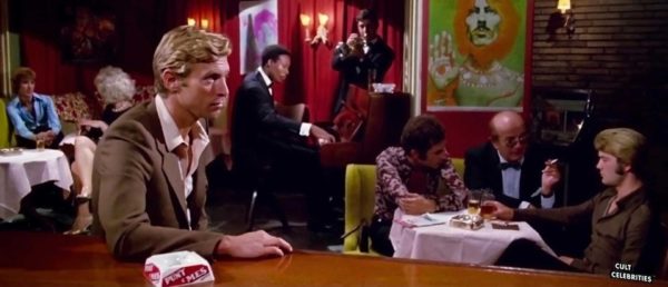 A blonde man sits at a table in a crowded gay bar