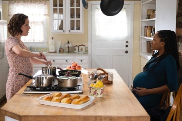 A white woman cooks breakfast for a seated pregnant black woman on the other side of the kitchen island
