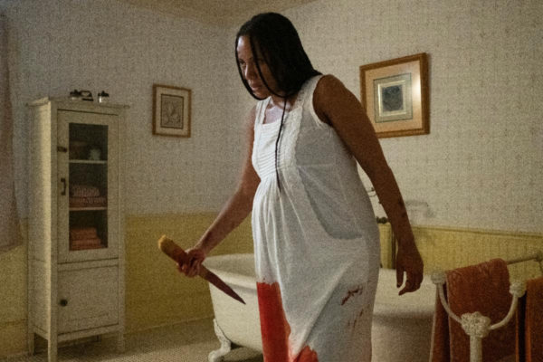 A pregnant woman in a bloody nightgown wields a stake in a bathroom