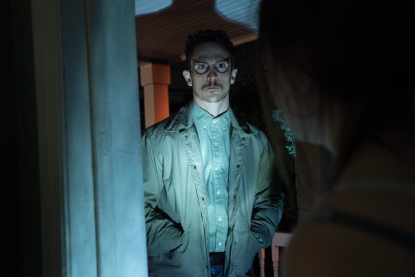 A bespectacled and mustachoed man stands in a doorway in the light of a flashlight