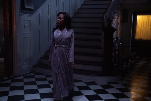 A woman in a nightgown stands at the base of a tall flight of stairs in the dark