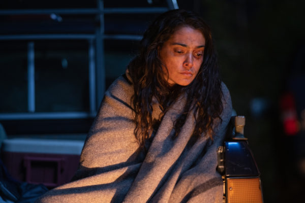 A woman with facial bruises sits in front of a campfire, wrapped in a blanket