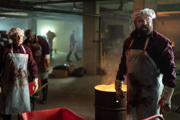 A large bearded man in an apron and gloves stands in front of a dumpster