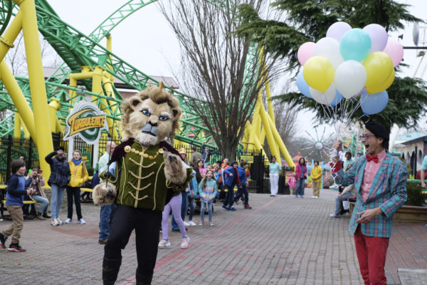 A man with balloons gazes at a another man in a prince outfit with a large grizzly mascot head