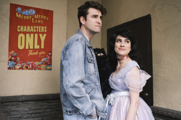 A tall dark haired man in glasses and a jean jacket stands next to a brunette in a princess costume