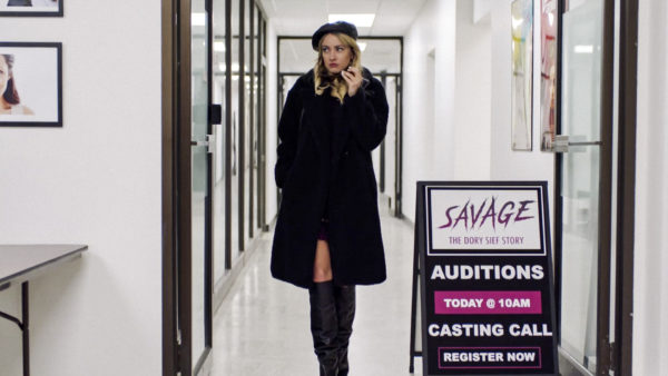 A stylish blonde dressed all in black stands next to a sign that reads SAVAGE auditions