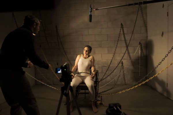 A woman dressed in white with a shaved head is tied to a chair in front of a camera