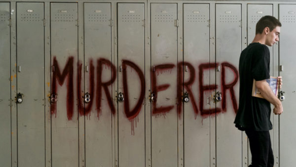 A teen boy walks past a row of lockers with the word MURDERER written in red paint