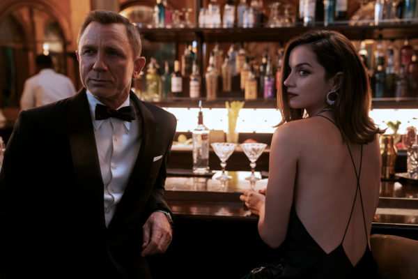A blonde man in a tux leans against a bar next to a brunette woman in a backless dress
