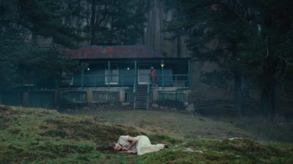 A woman in a white nightgown lays on the ground in front of a cottage in the woods