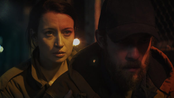 A woman stands behind a man wearing a baseball cap with his head down, illuminated from behind