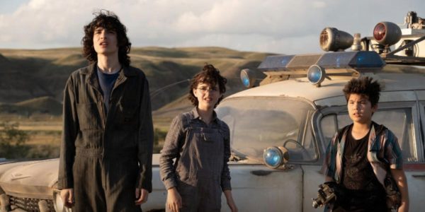 A male teenager (L) and two pre-teens - a girl and an asian boy - stand in front of a dusty car