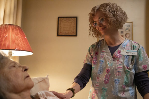 A female nurse in glasses and frizzy hair comforts an elderly woman in bed