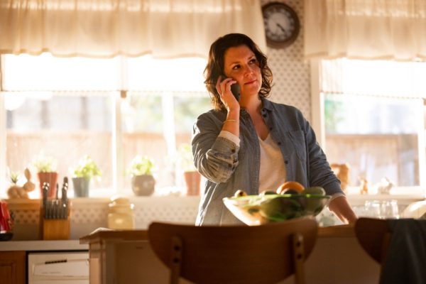 A brunette woman standing in a kitchen listens on the phone