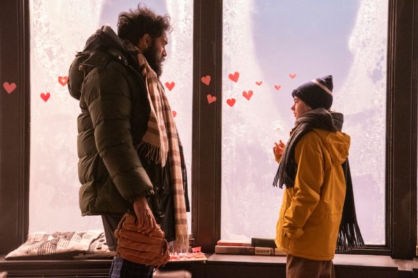 A man and a little girl in winter coats stand in front of a window covered in paper hearts