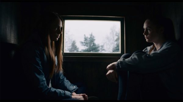 A red headed woman sits facing her double in a tight enclosed room