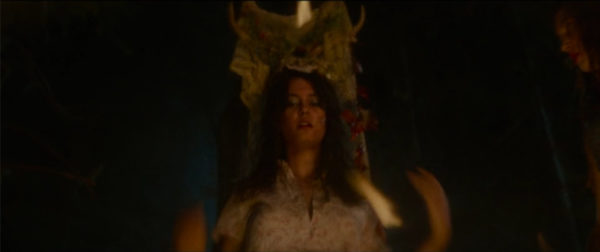 A brunette teen girl standing in front of a fire wearing a ceremonial headdress