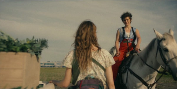 A young woman on horseback speaks to a woman standing with her back to us