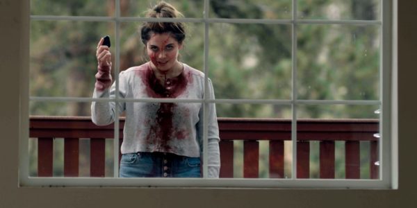 A woman in a bloody shirt holds a knife in a window