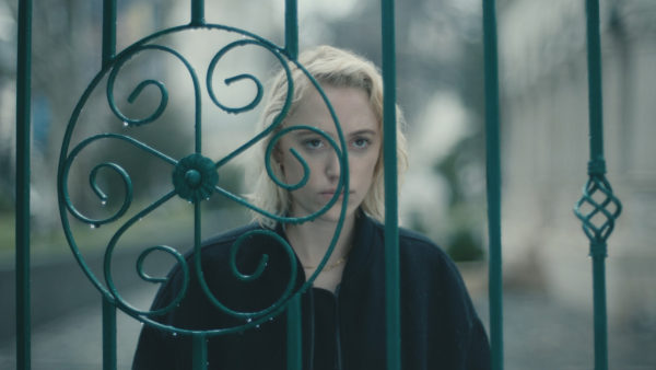 A blonde woman stares throw a wrought iron gate