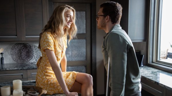 A blonde woman sits on the kitchen counter in front of a man on crutches who is wearing glasses 