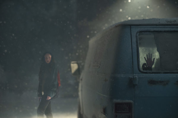 A young woman stands in the snow next to a van with a person banging on the window