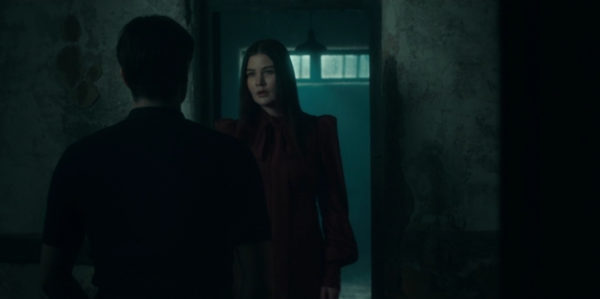 A dark haired woman standing in a blue-lit door, talking to a man facing away from us
