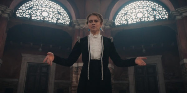 A blonde woman in a velvet coat stands with arms outstretched