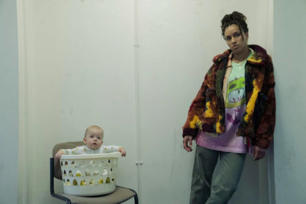 A woman leans against a wall next to a baby in a laundry bin