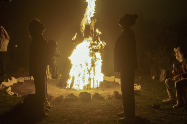 A old woman (L) holding a baby faces a young woman (R) in front of a bonfire