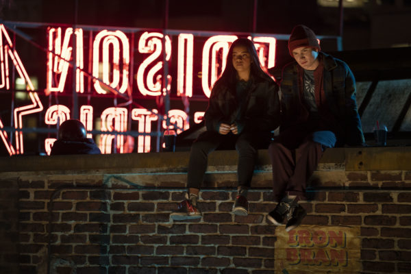 2 teenagers sit on the ledge of a roof in front of a neon sign