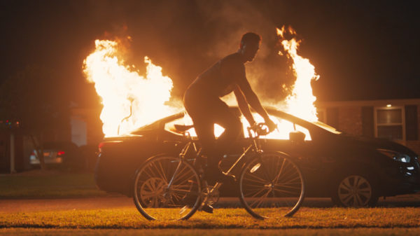 A man on a bike in front of a car on fire