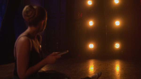 A ballerina looks at a masked man on the edge of the stage