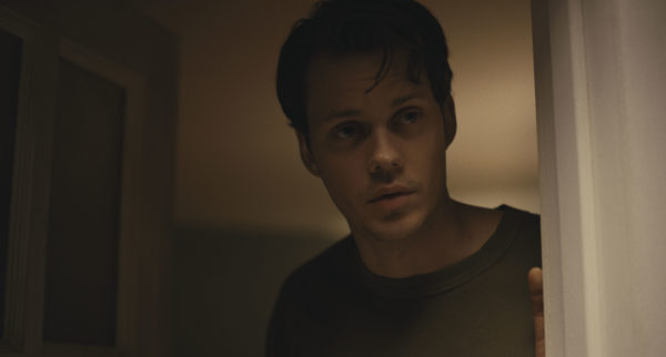 A man (Bill Skarsgård) lit in shadows, looks out an open door