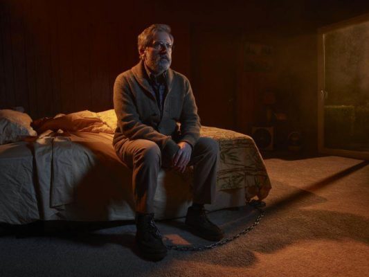 A man (Steve Carell) sits on a bed with a leg shackled in chains