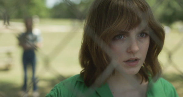 Mckenna Grace as Jan Broberg, staring through a chain link fence