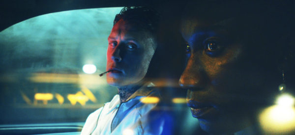 A white man with a cigarette (left) watches a black man (r) driving