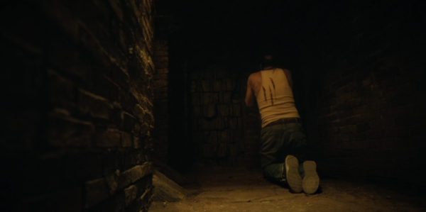 Uncle George (Boris McIver) kneeling, facing away from the camera, with bloody marks on his back
