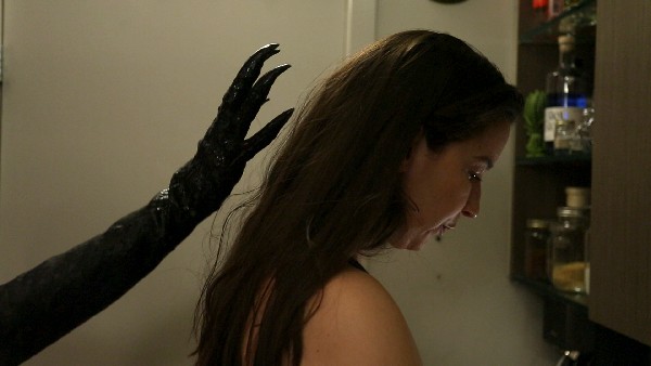 A dark, taloned hand reaches towards a woman (R) facing away from it