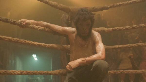 A tank top wearing man wearing a monkey mask poses against boxing rings