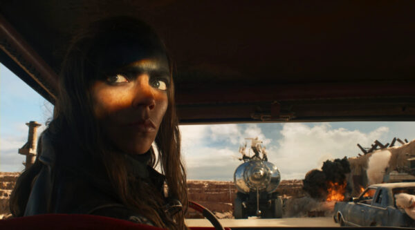 A close-up of Furiosa (Anya Taylor-Joy)'s face as she sits in the driver's seat of a vehicle with fiery chaos behind her