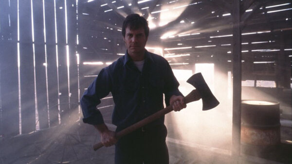 A man with an axe stands in a barn with a bright light illuminating him from behind