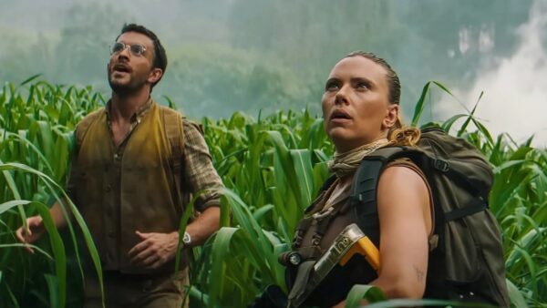 A dark haired man in slutty glasses stares in awe next to a blonde woman in a tank top, holding a gun, amidst tall ferns