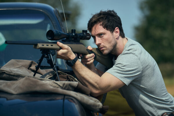 A muscley white man takes aim through a telescopic rifle on the hood of a car